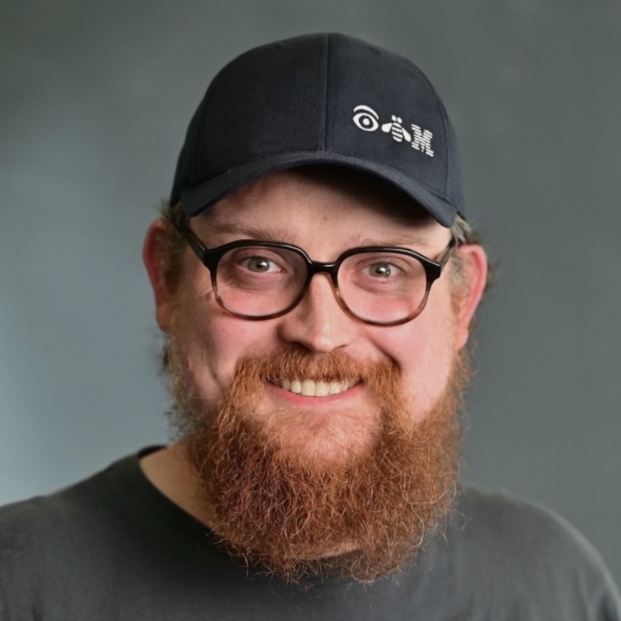 A smiling man with glasses and a beard wearing an IBM-branded baseball cap.